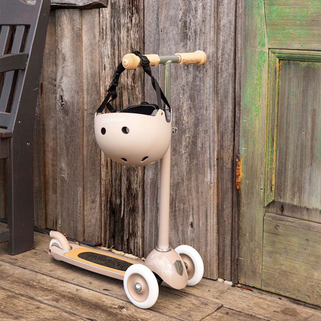 Banwood kinderstep in beige met een bijpassende helm hangend aan het stuur, staand tegen een houten muur.