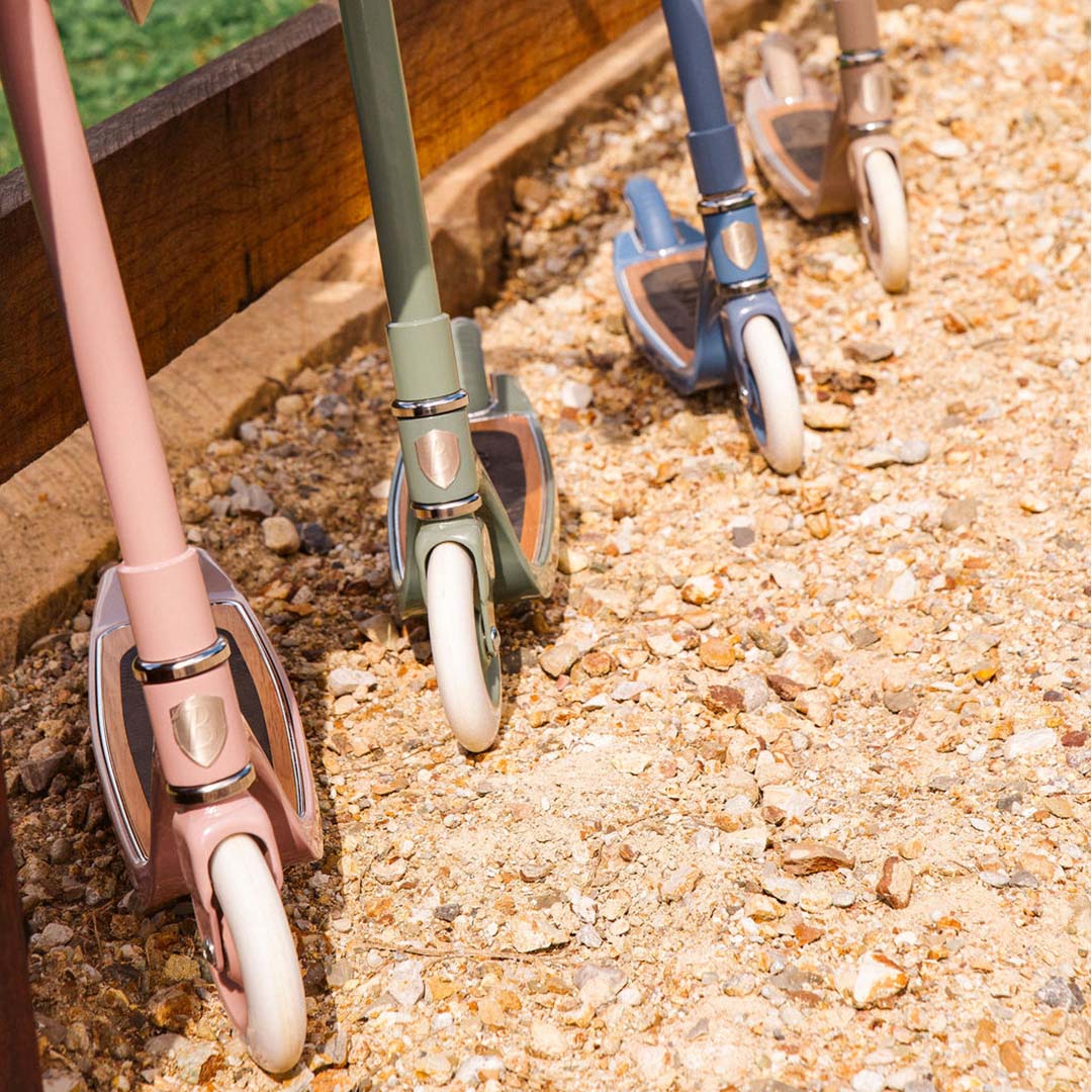 Drie Banwood kindersteps in verschillende kleuren staan op een grindpad naast een houten hek.