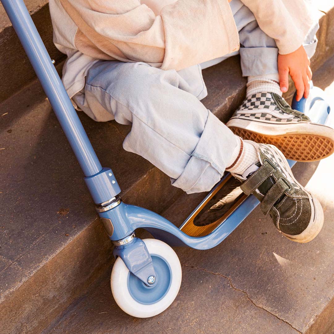 Detail van een kind dat naast een lichtblauwe Banwood kinderstep op stenen trappen zit.