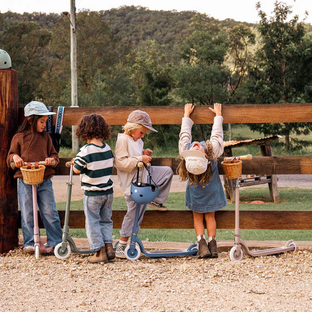 Vier kinderen staan met verschillende Banwood kindersteps bij een houten hek in een landelijke omgeving.