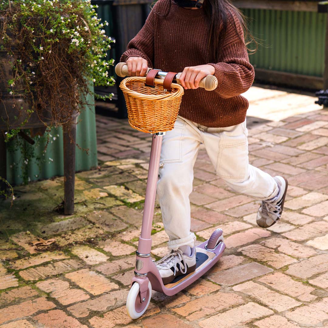 Een kind rijdt op de Banwood kinderstep Maxi in dusty roze met rieten mandje op een bakstenen pad.