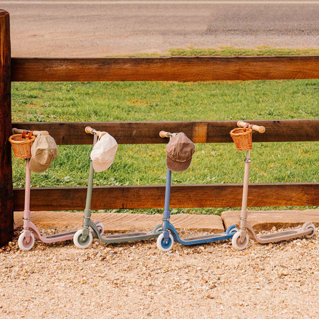 Vier Banwood kindersteps in verschillende kleuren, met petjes aan het stuur, langs een houten hek.
