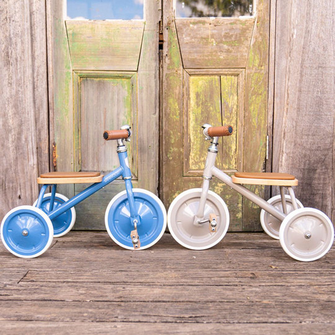 Twee Banwood driewielers, een blauwe en een beige, staan naast elkaar voor een houten deur.