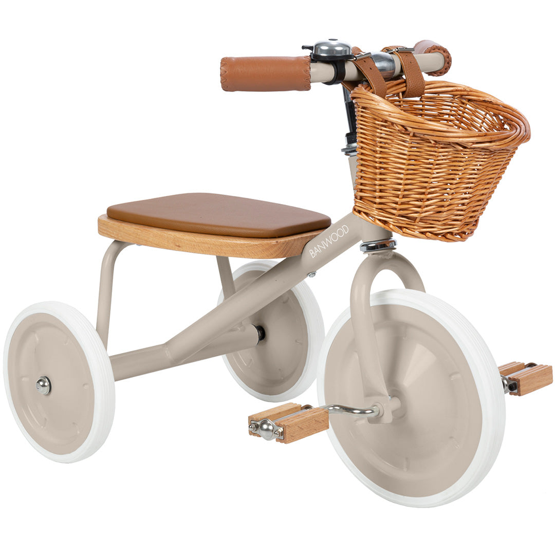 Banwood beige driewieler met bruin zadel, houten pedalen en rieten mandje, schuin van voren gefotografeerd.