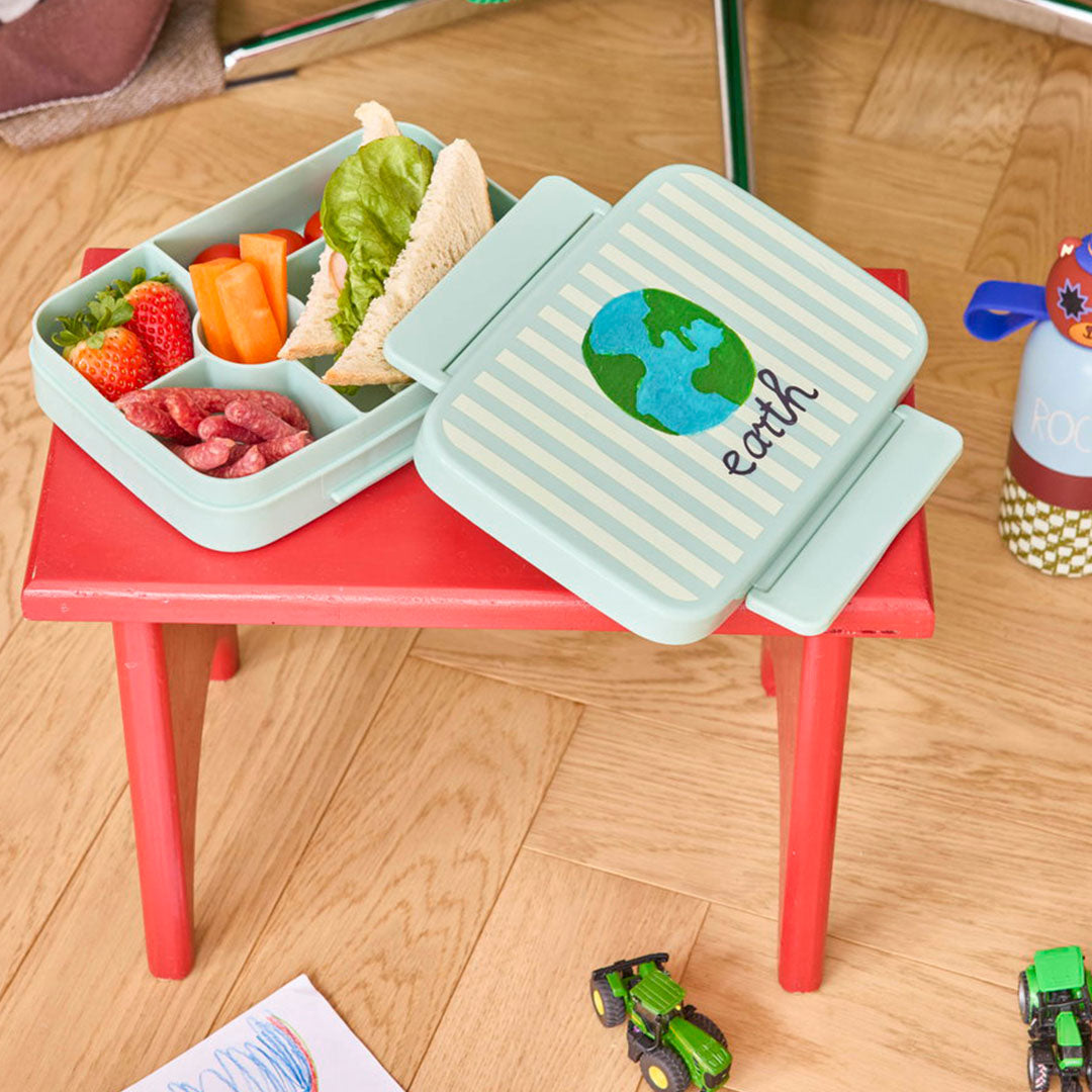 Rice bento broodtrommel met aarde-illustratie, gevuld met fruit, groenten en een boterham op een rood tafeltje.