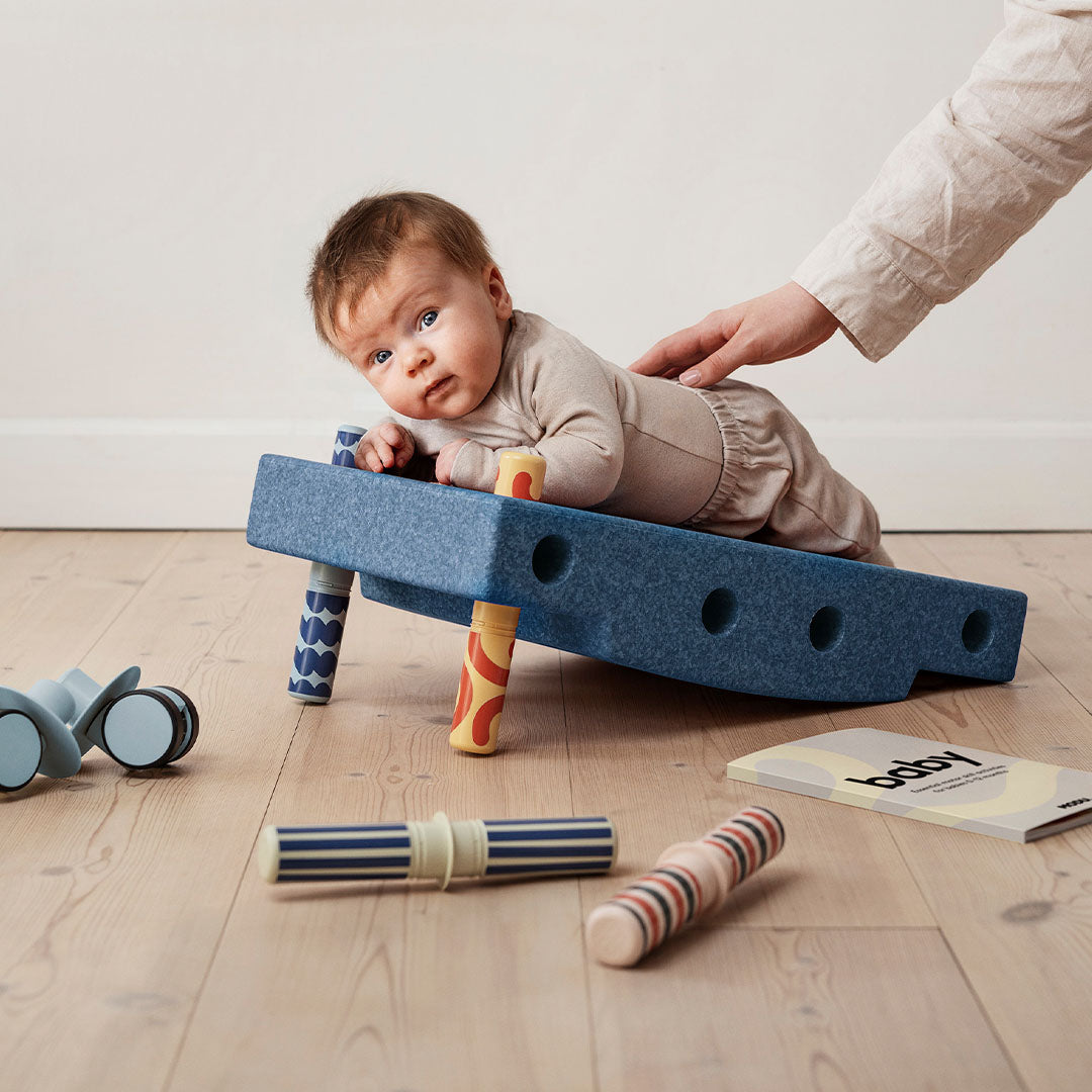 Een baby ligt op de buik op een blauwe Modu babyactiviteitenset, met een hand die de rug ondersteunt.