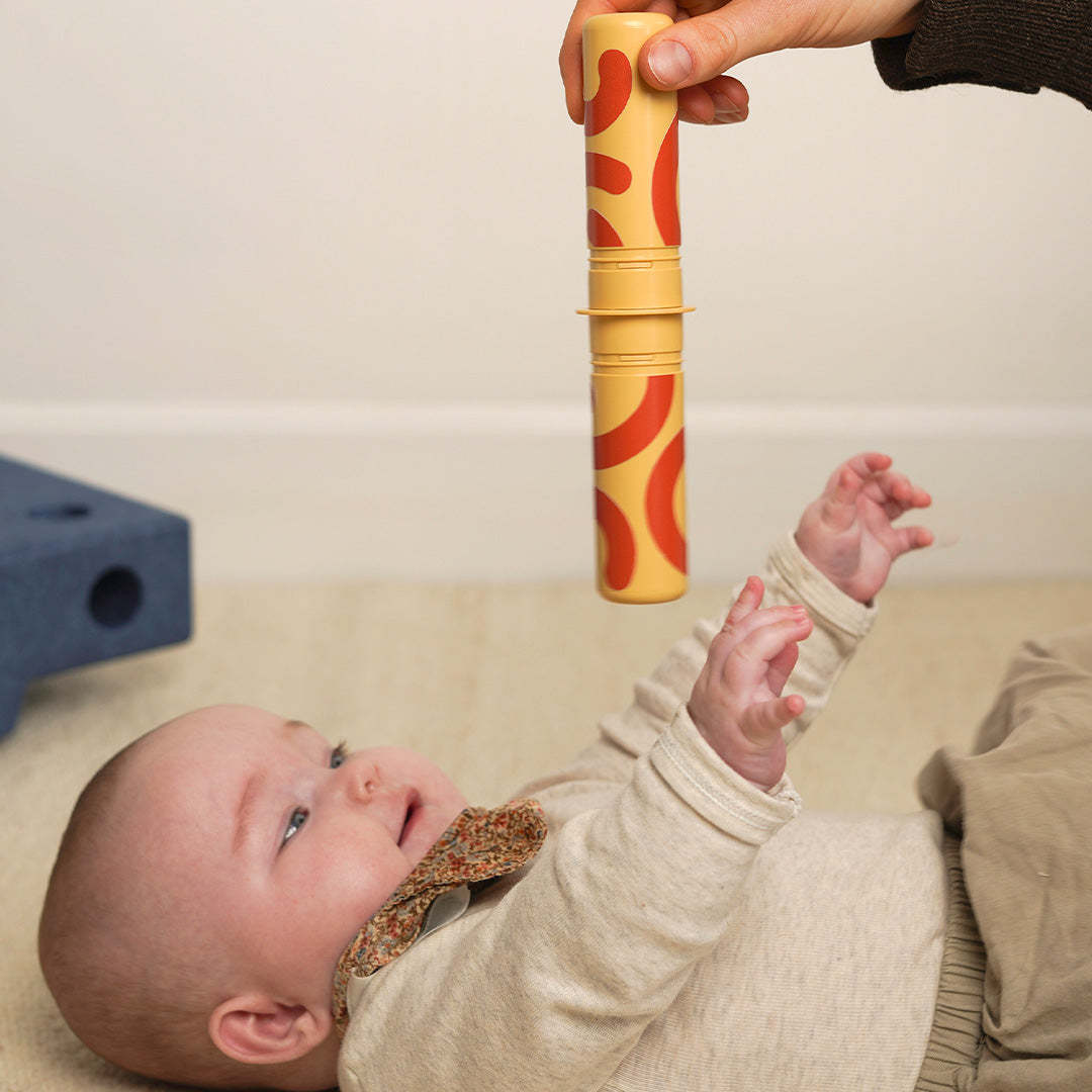 Een baby ligt op de rug en kijkt naar een gele rammelaar met rode patronen die boven hem wordt gehouden.