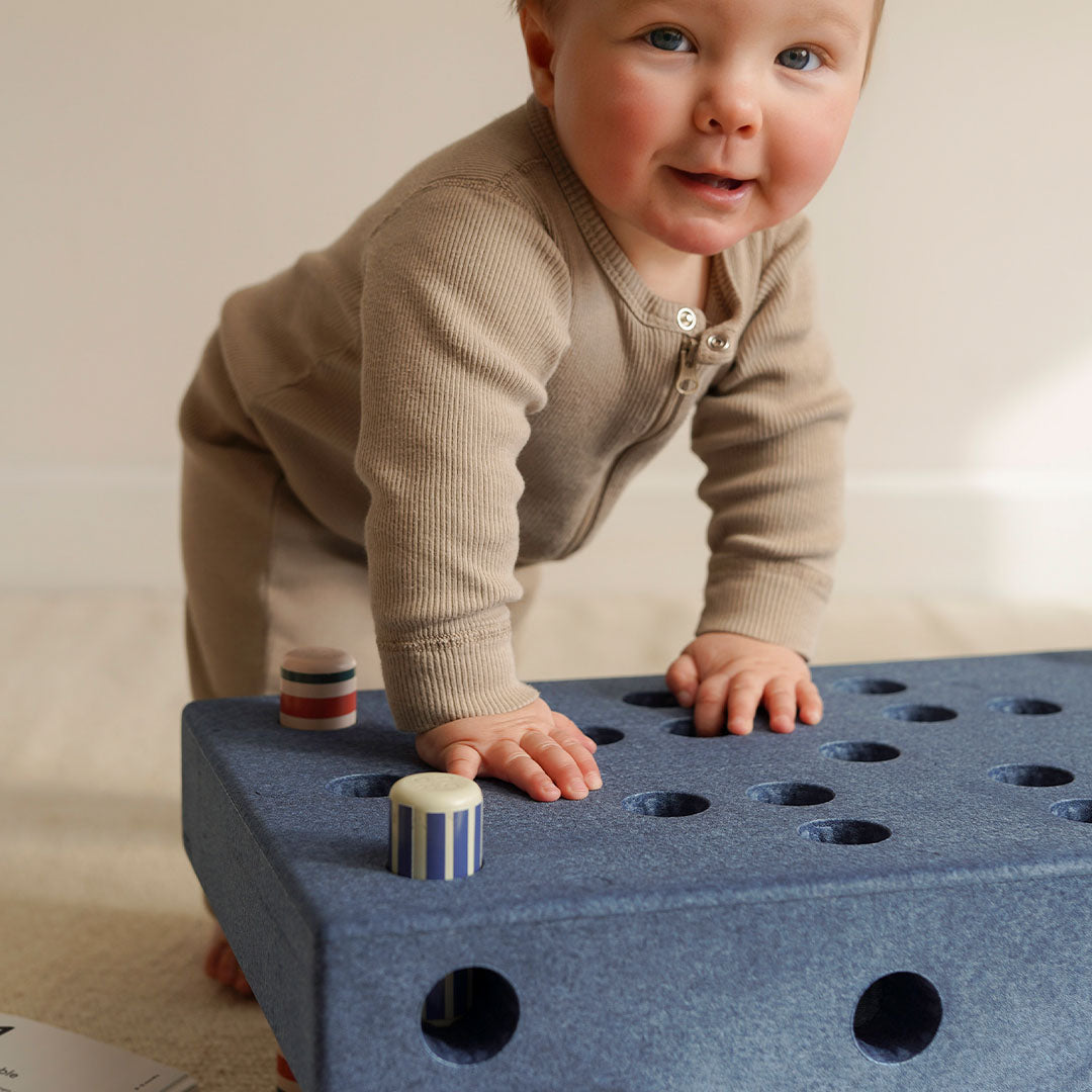 Een baby kruipt op handen en knieën naast een blauwe Modu babyactiviteitenset met gekleurde pinnen.