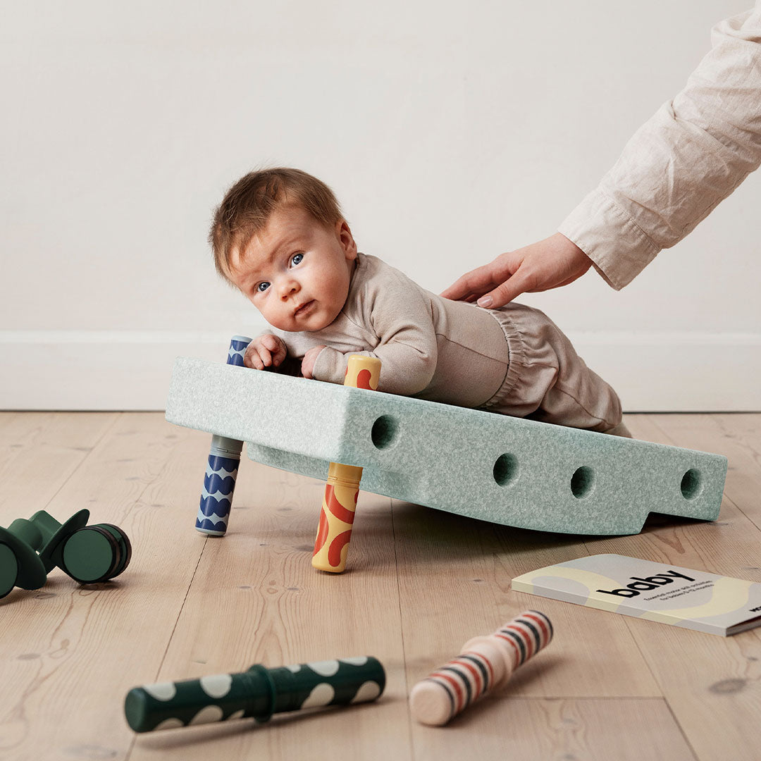 Een baby ligt op de buik op een groene Modu schuim activiteitenset, met een hand die de rug ondersteunt.
