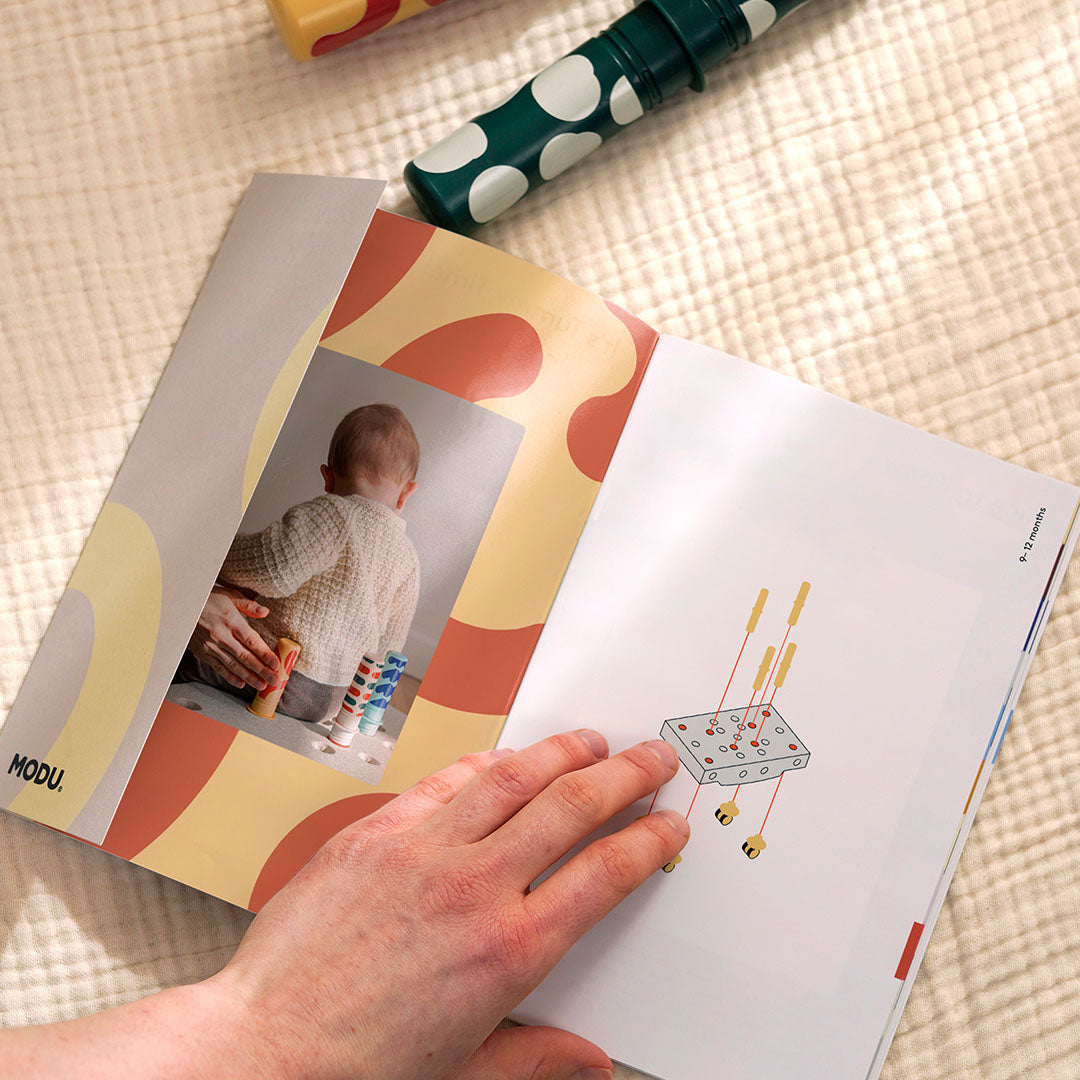 Een hand bladert door het Modu instructieboekje met montage-illustraties en een foto van een spelende baby.