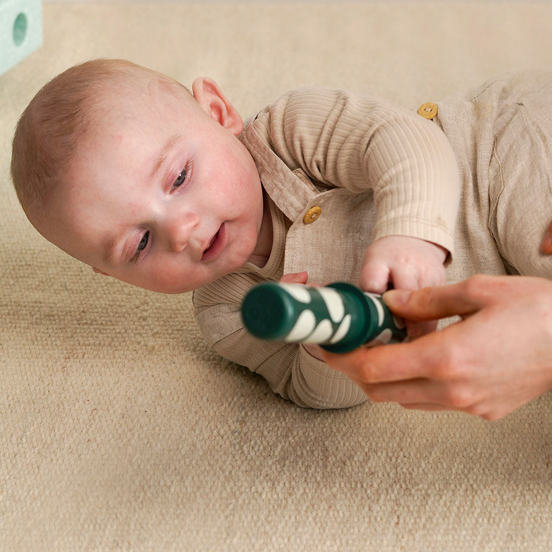 Een baby op een lichte ondergrond reikt naar een groene rammelaar met witte patronen die door een hand wordt vastgehouden.