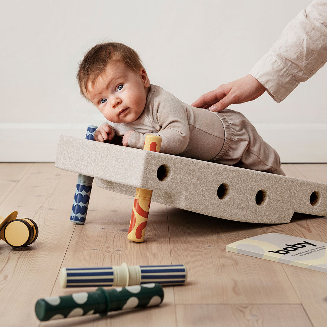 Een baby ligt op zijn buik op een modu schuimrubberblok dat is opgebouwd als wiebelend speelgoed, ondersteund door een hand.