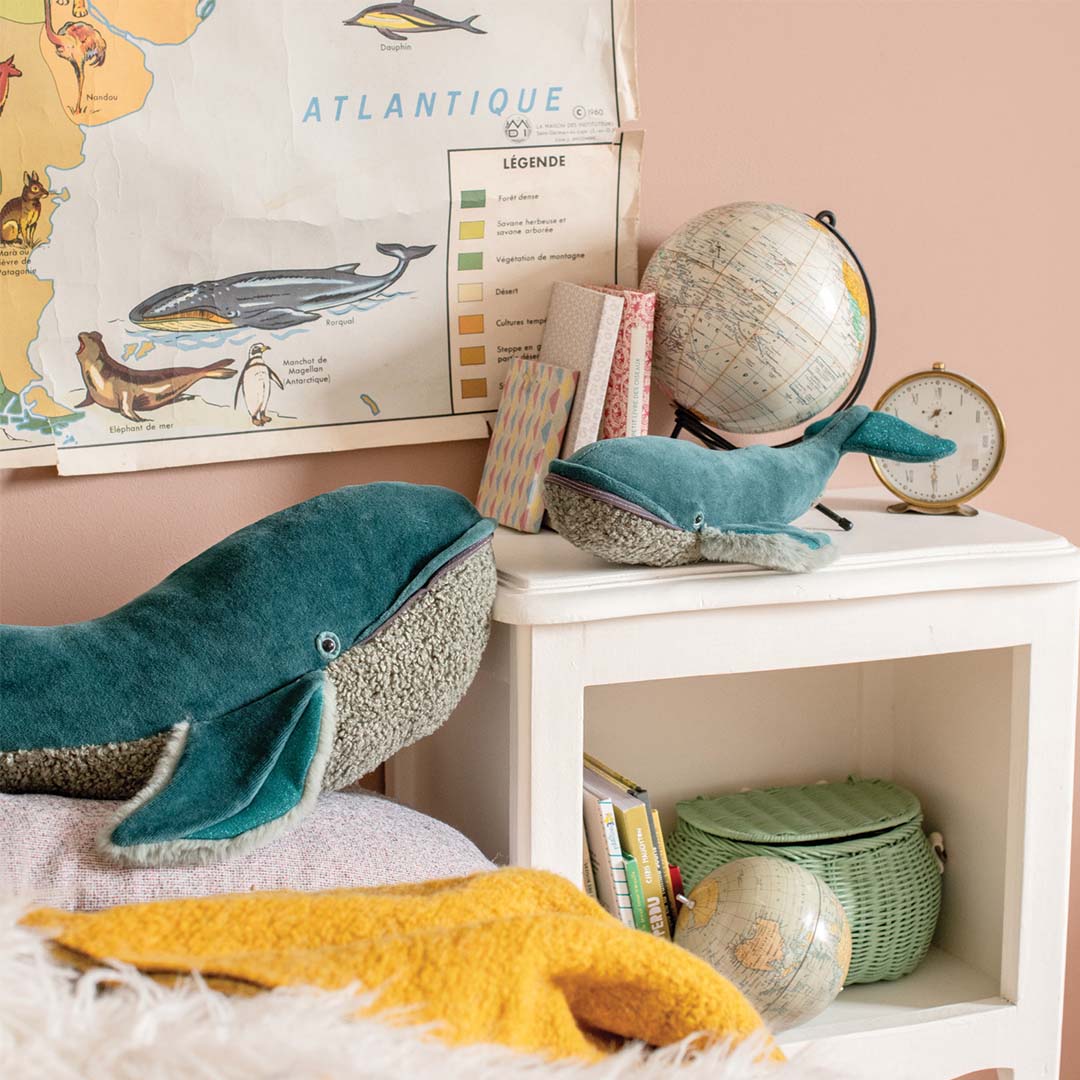 Twee Moulin Roty knuffelwalvissen op een bed en een nachtkastje in een kinderkamer met een wereldkaart.