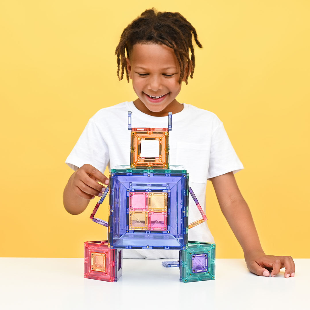 Een lachende jongen bouwt een robot met Connetix magnetische tegels in pastelkleuren.
