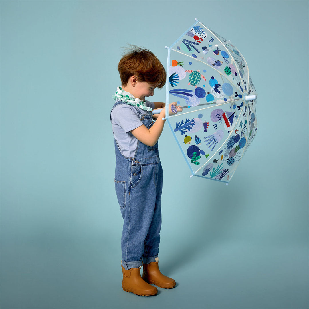 Een kind in een tuinbroek houdt de Djeco kinderparaplu met zeedierenpatroon vast.