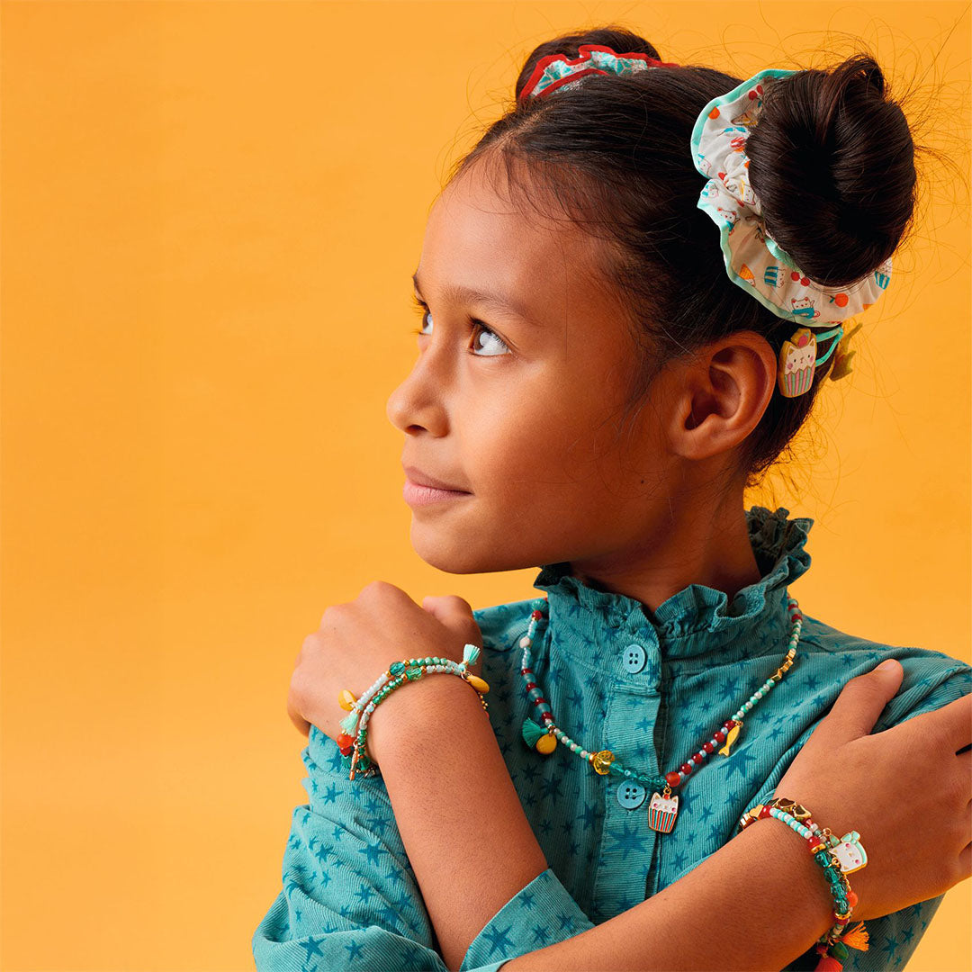 Een kind draagt de Djeco kinderketting en bijpassende kralenarmbanden, met haar in knotjes en scrunchies, tegen een gele achtergrond.