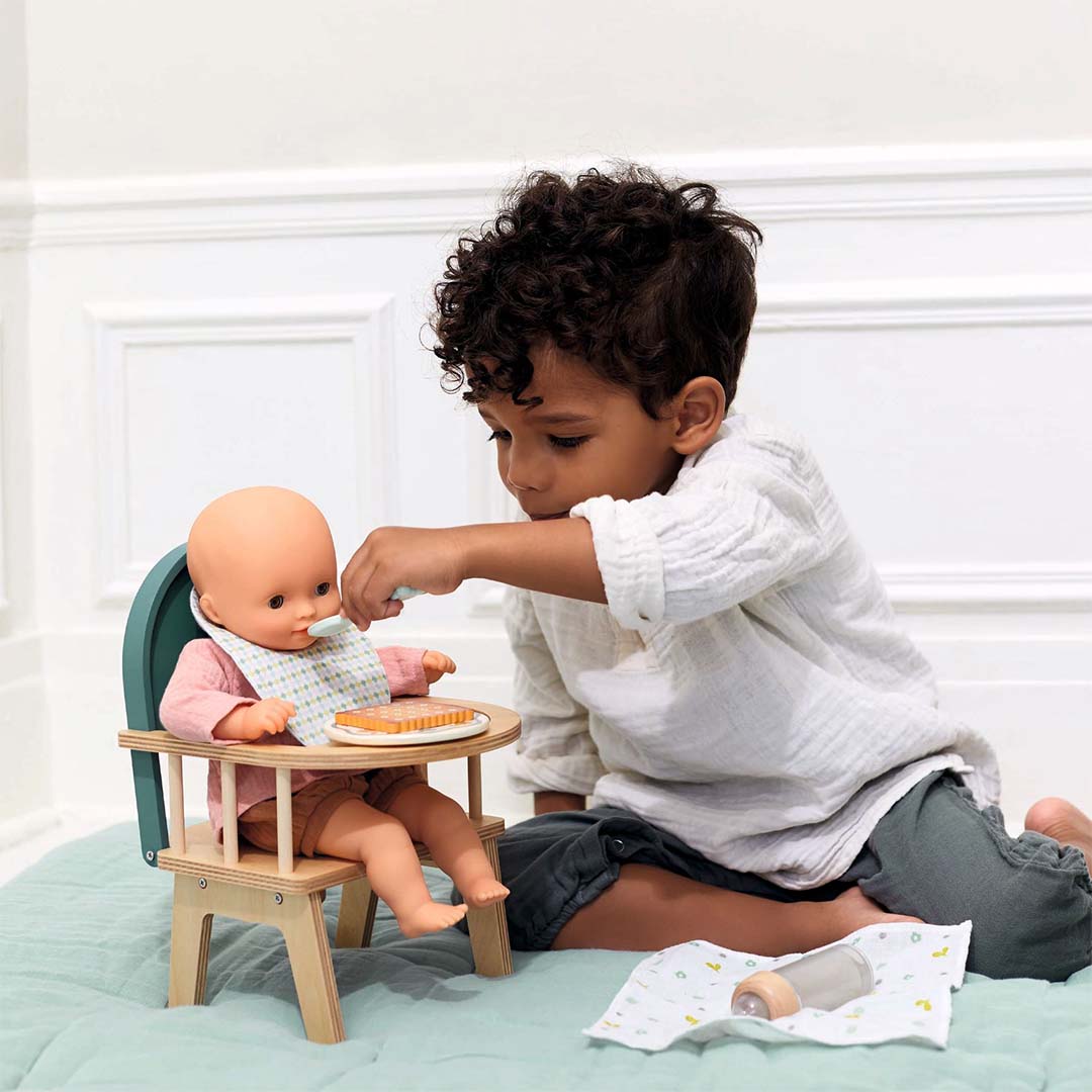 Een kind voedt een pop in een houten kinderstoel met een lepel en koekje uit de Djeco Pomea poppenvoedingsset.