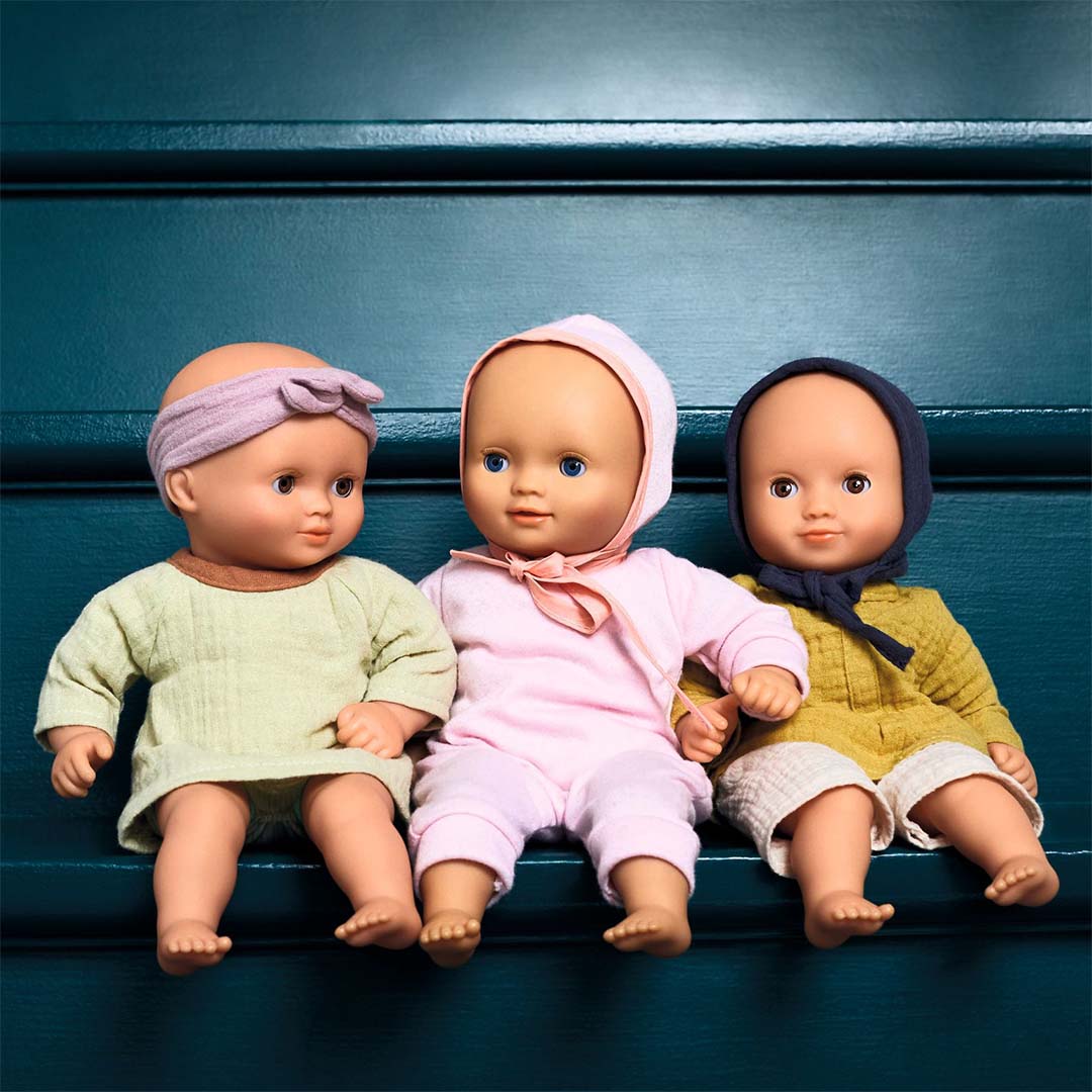 Drie Djeco babypoppen Pomea in verschillende outfits, zittend naast elkaar op een donkerblauwe ondergrond.