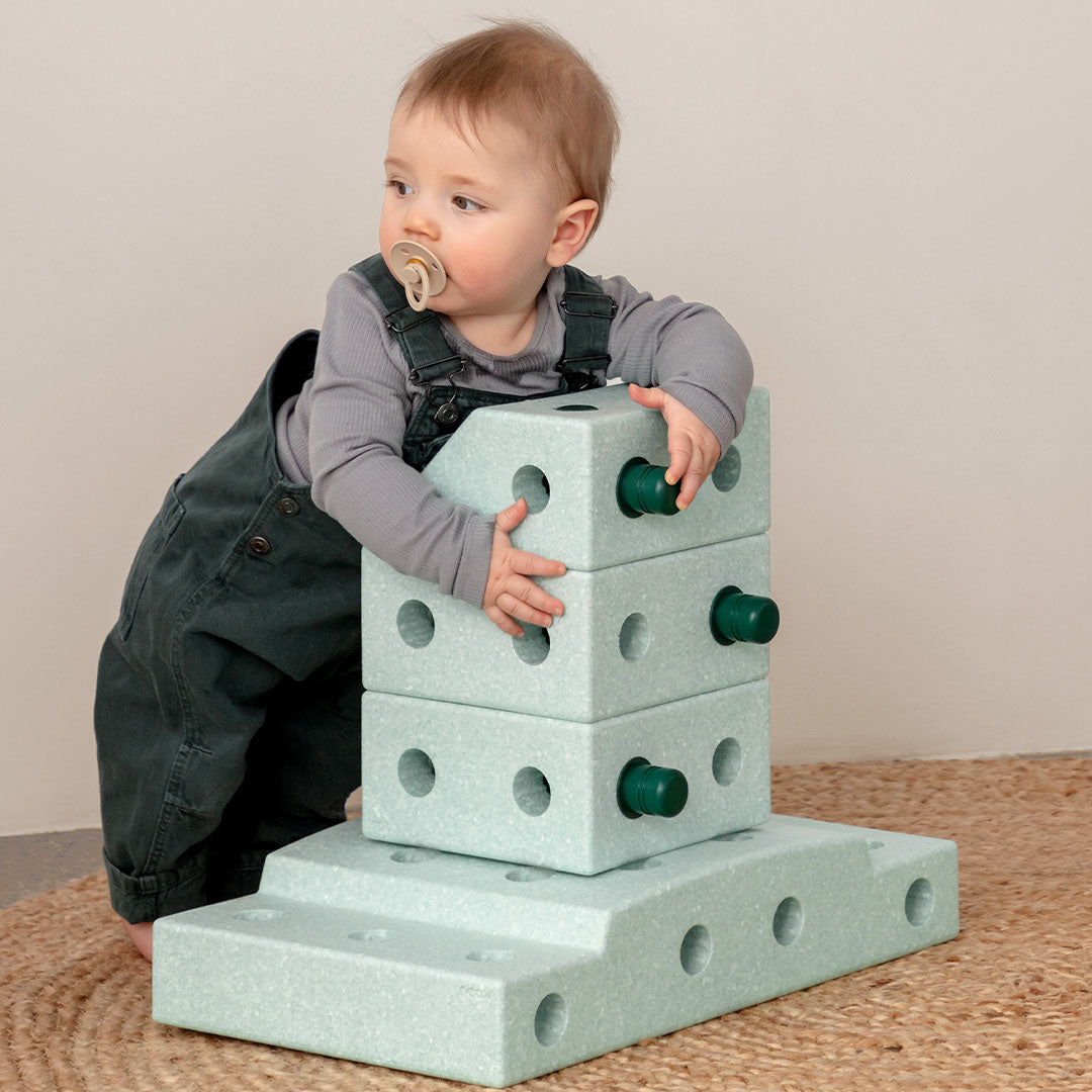Een baby staat naast een toren van lichtgroene Modu schuimrubberblokken met groene verbindingsstukken.