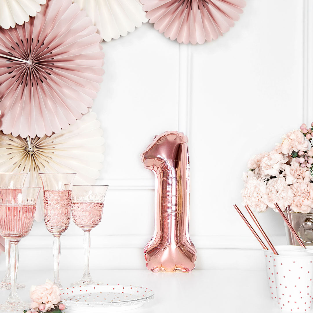 Rosegouden cijferballon 1 op een witte tafel met roze feestdecoraties.