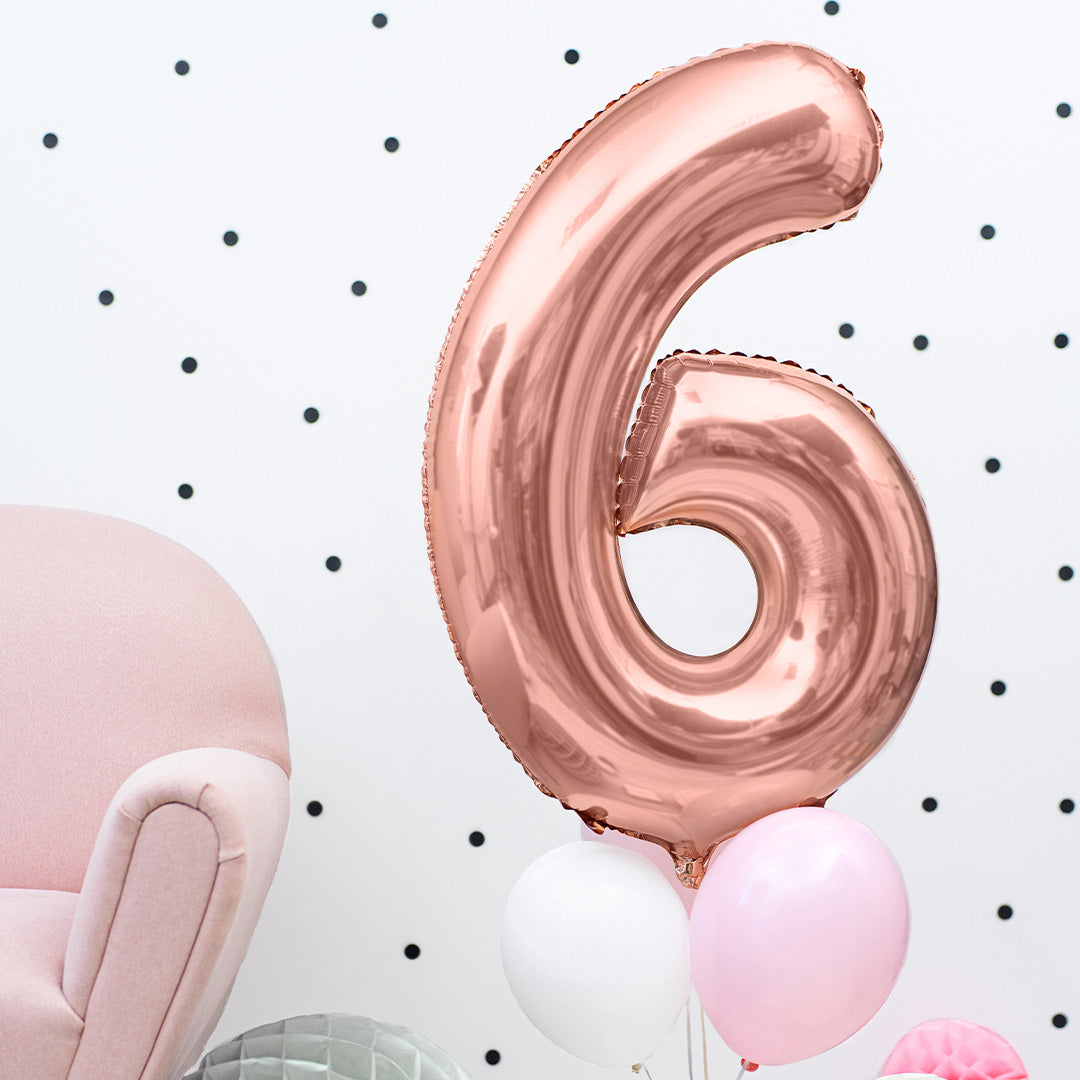 Partydeco roségouden cijferballon 6 zwevend met een witte en roze ballon in een kamer met stippenbehang en een roze fauteuil.