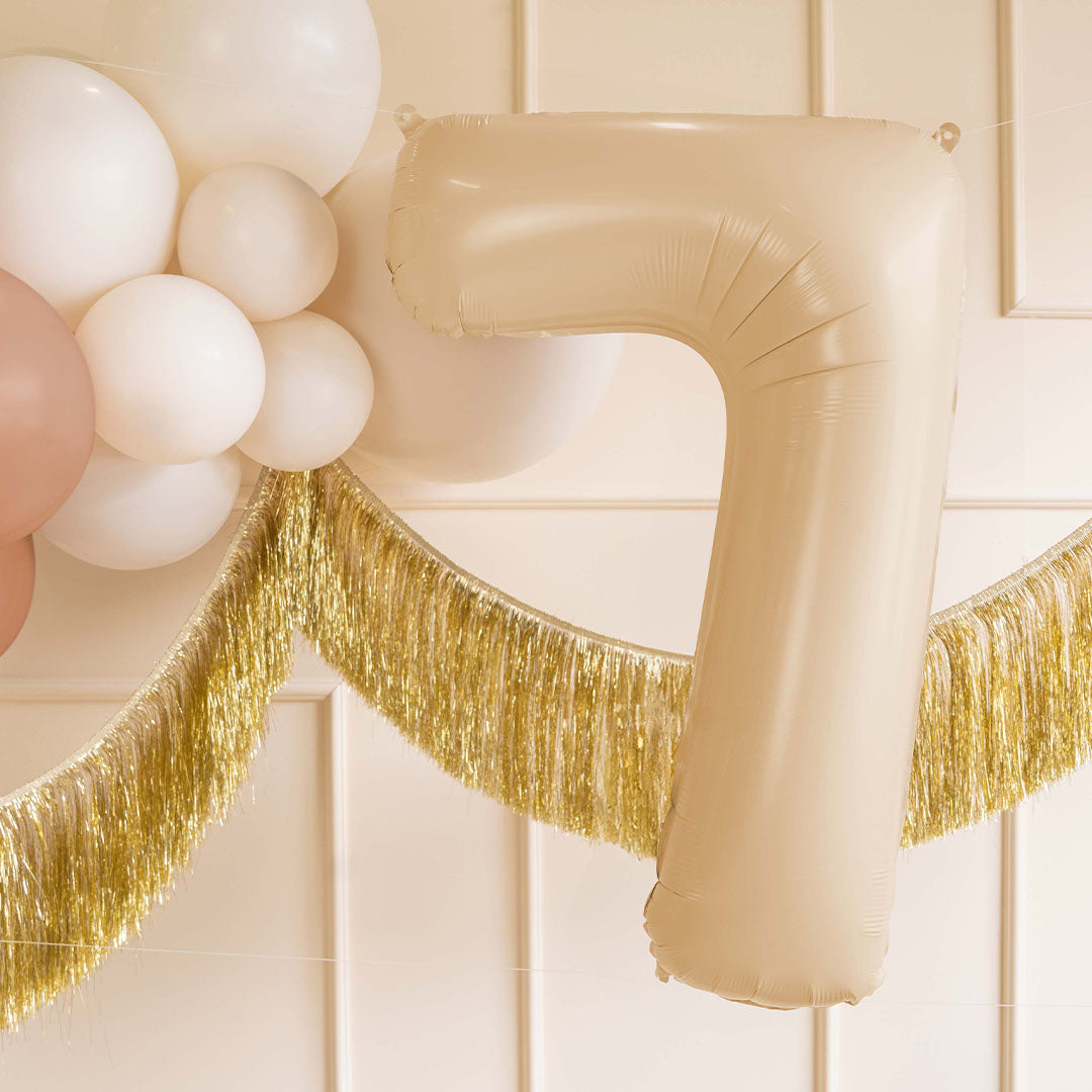 Beige cijfer 7 ballon hangend tussen witte ballonnen en een gouden franjeslinger als feestdecoratie.