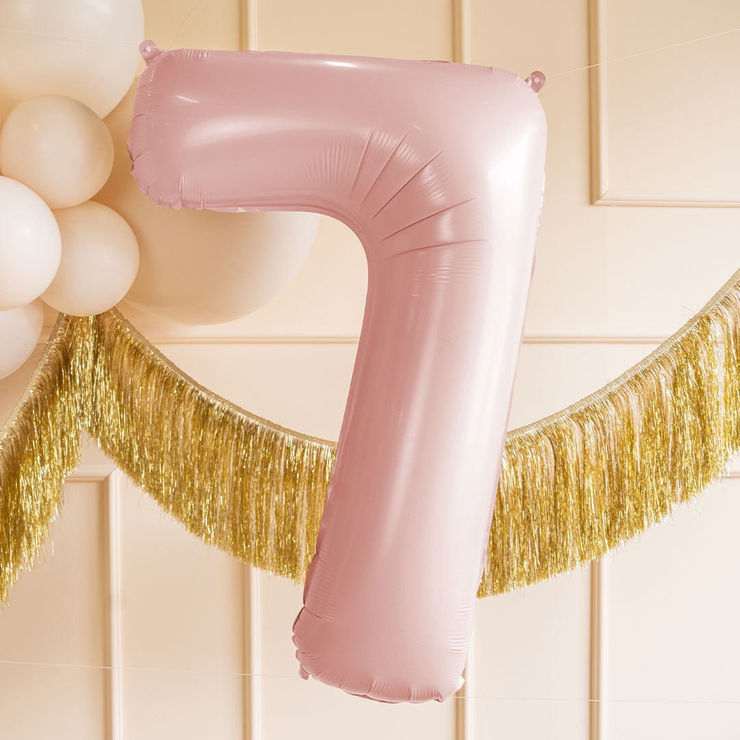 Lichtroze cijferballon 7 als feestdecoratie met witte ballonnen en een gouden slinger op de achtergrond.