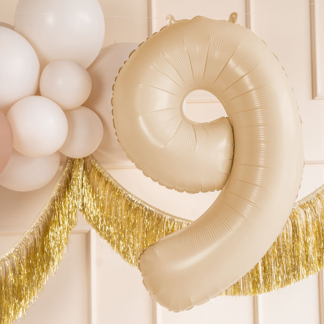Partydeco beige cijferballon 9 hangend tussen witte ballonnen en een gouden slinger als feestdecoratie.