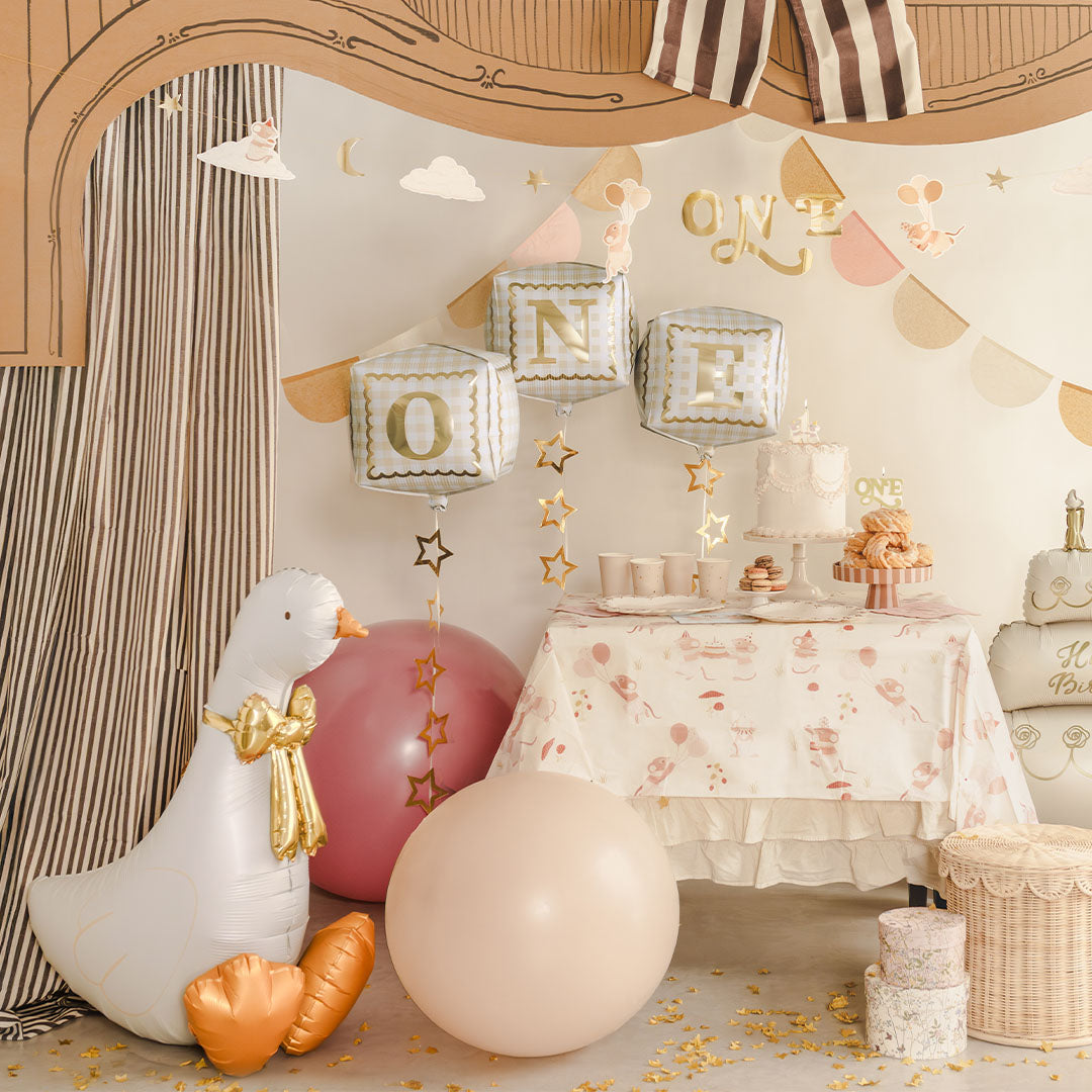 Een witte folieballon in de vorm van een gans staat in een feestelijke setting met 'ONE' ballonnen en een verjaardagstaart.