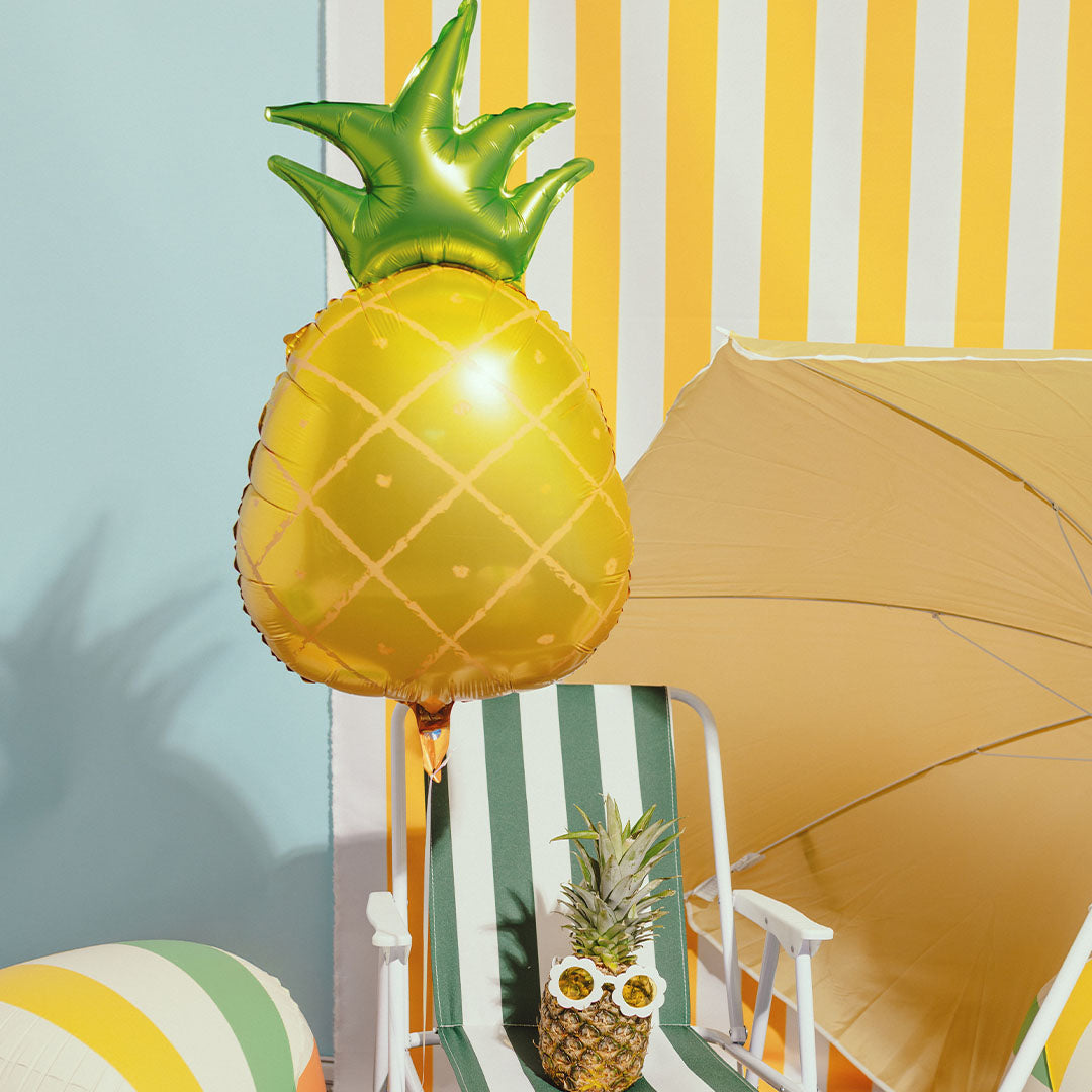 Een PartyDeco ananas folieballon zweeft boven een strandstoel met een echte ananas.