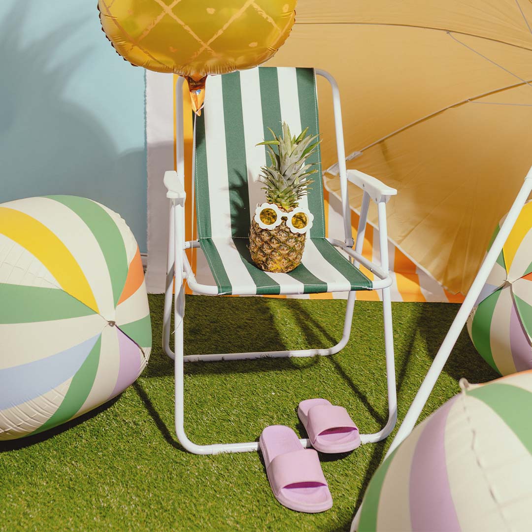Een PartyDeco gestreepte folieballon ligt op het gras naast een strandstoel met een ananas en slippers.
