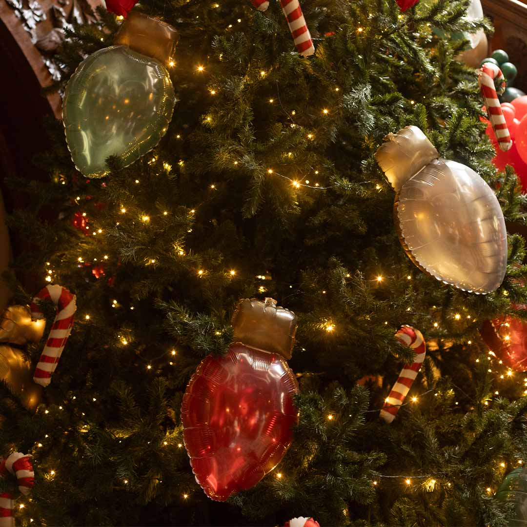 partydeco folieballonnen in de vorm van kerstlampjes hangend in een verlichte kerstboom.