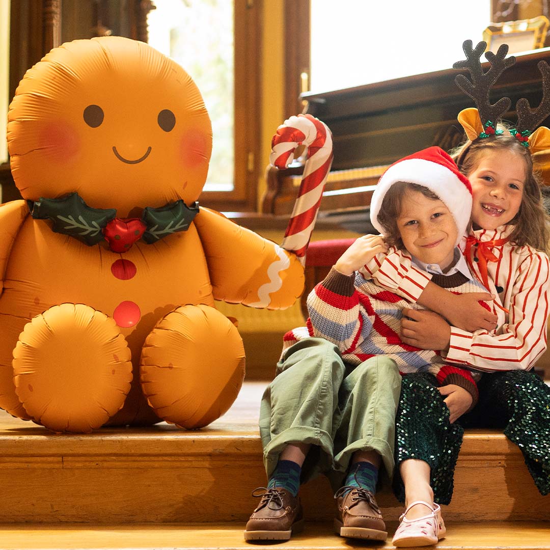 Twee lachende kinderen met kerstaccessoires zitten naast een grote opblaasbare gingerbread man folieballon.