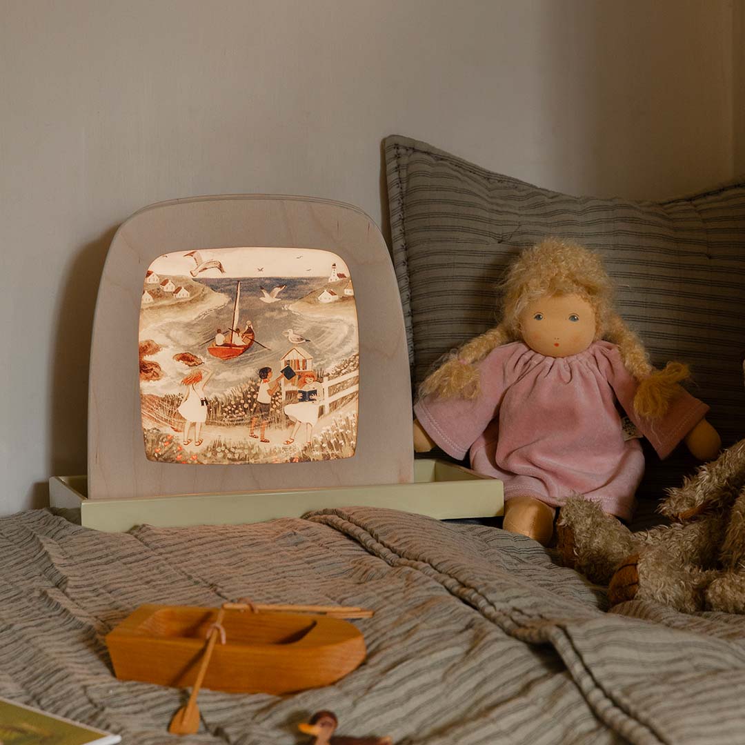 De Toverlux toverplaat 'aan de zee' verlicht in een toverlamp op een bed, naast een pop en een houten boot.