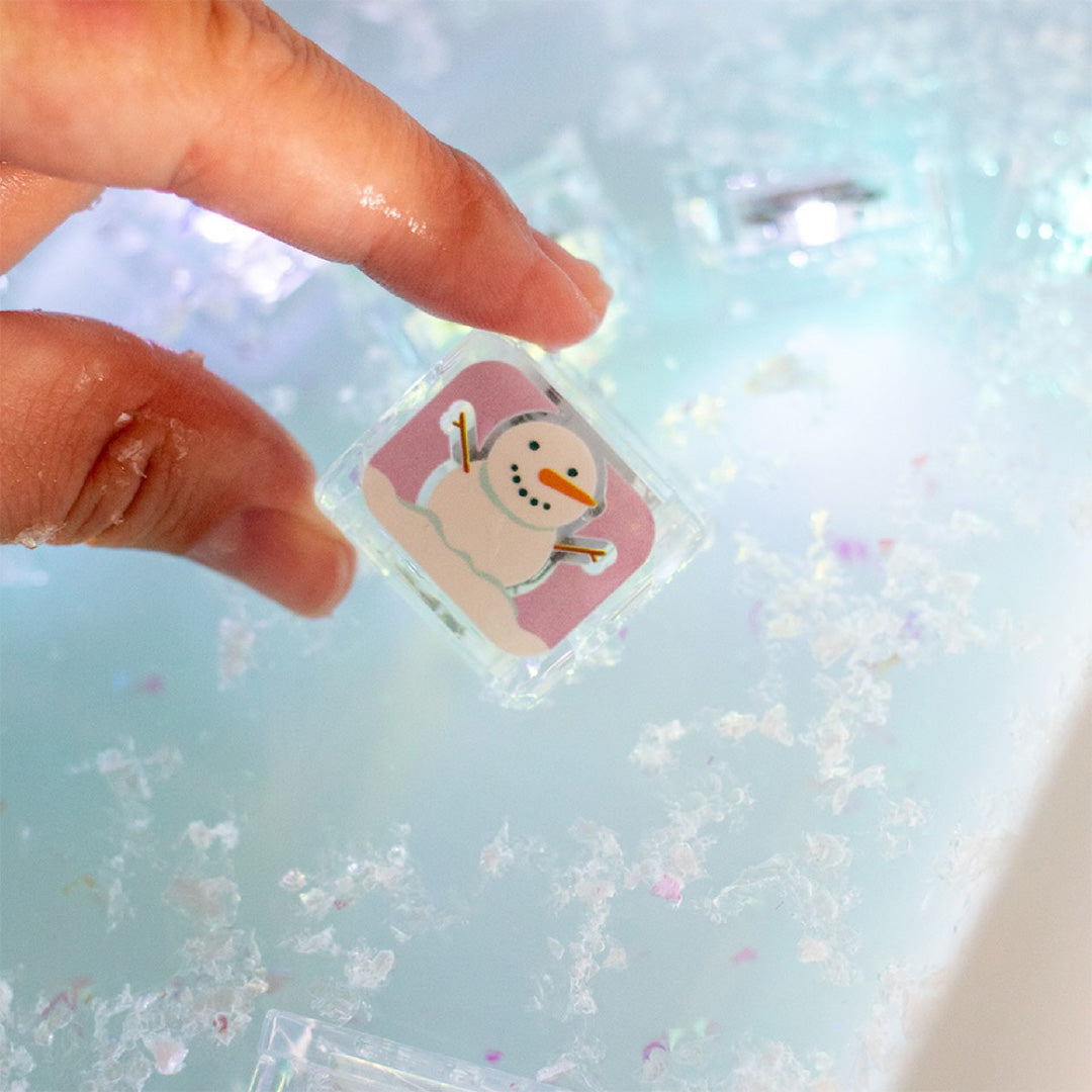 Een hand houdt een oplichtend glo pals badspeelgoedblokje vast met een smeltende sneeuwpop in water.