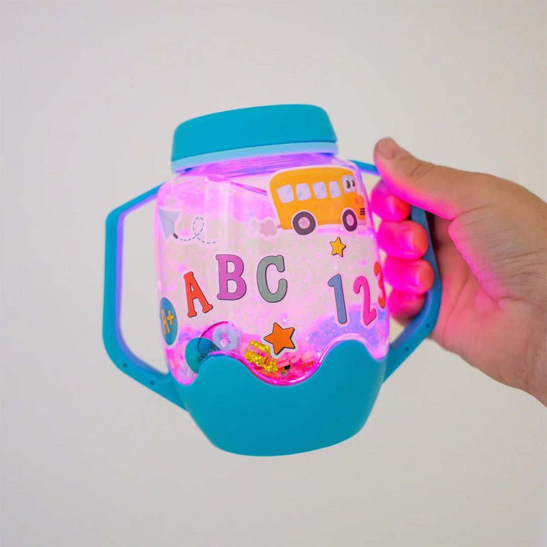Een hand houdt de Glo Pals DIY Sensory Play Jar met ABC thema vast, die roze oplicht en gevuld is met glitter en stickers.