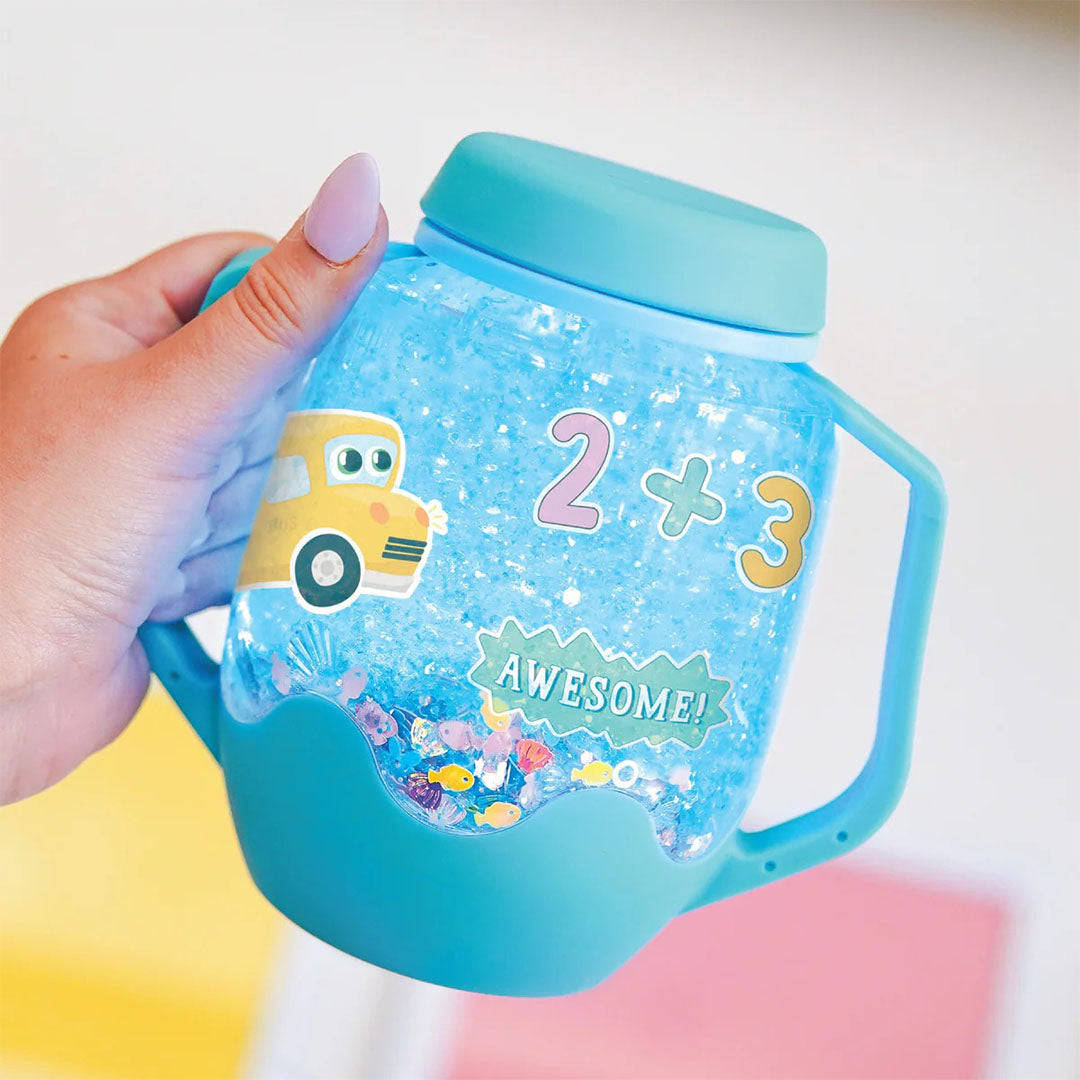 Een hand houdt de Glo Pals DIY Sensory Play Jar vast, die lichtblauw oplicht en gevuld is met blauwe glitter en stickers met een bus en cijfers.