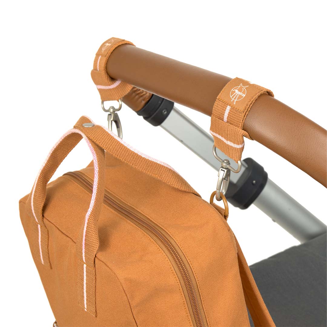 De Lässig caramel luiertas rugzak hangt met riemen aan de duwstang van een kinderwagen.