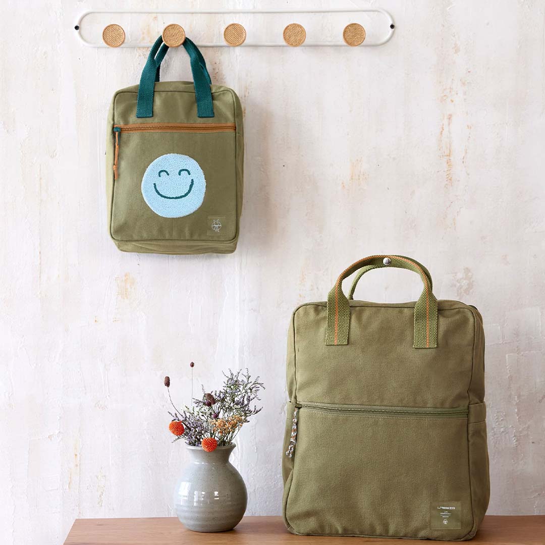 De Lässig kleuterrugzak met smiley hangt aan een haak naast een grotere bijpassende rugzak op een houten oppervlak.