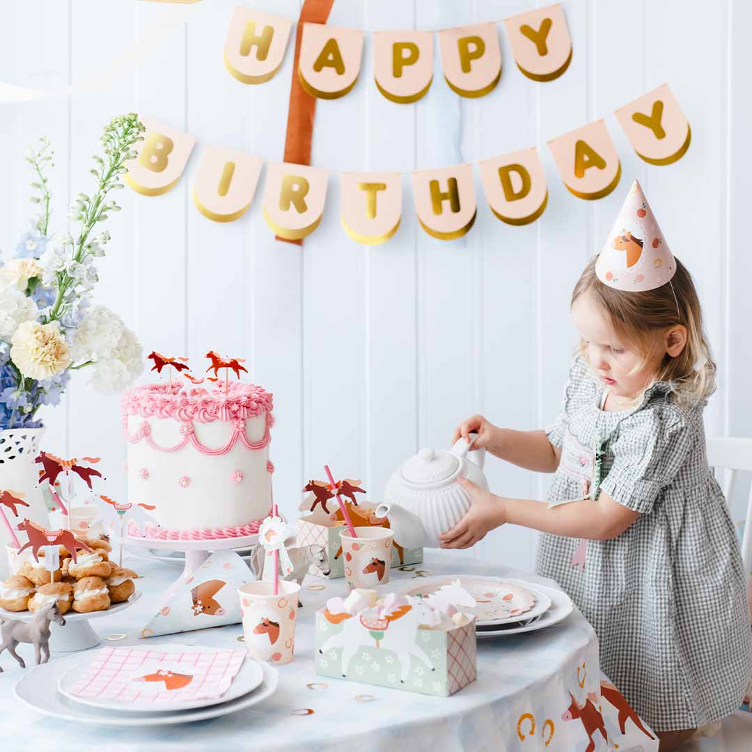 Een kind viert een verjaardagsfeestje met een PartyDeco roze en gouden 'Happy Birthday' slinger op de achtergrond.