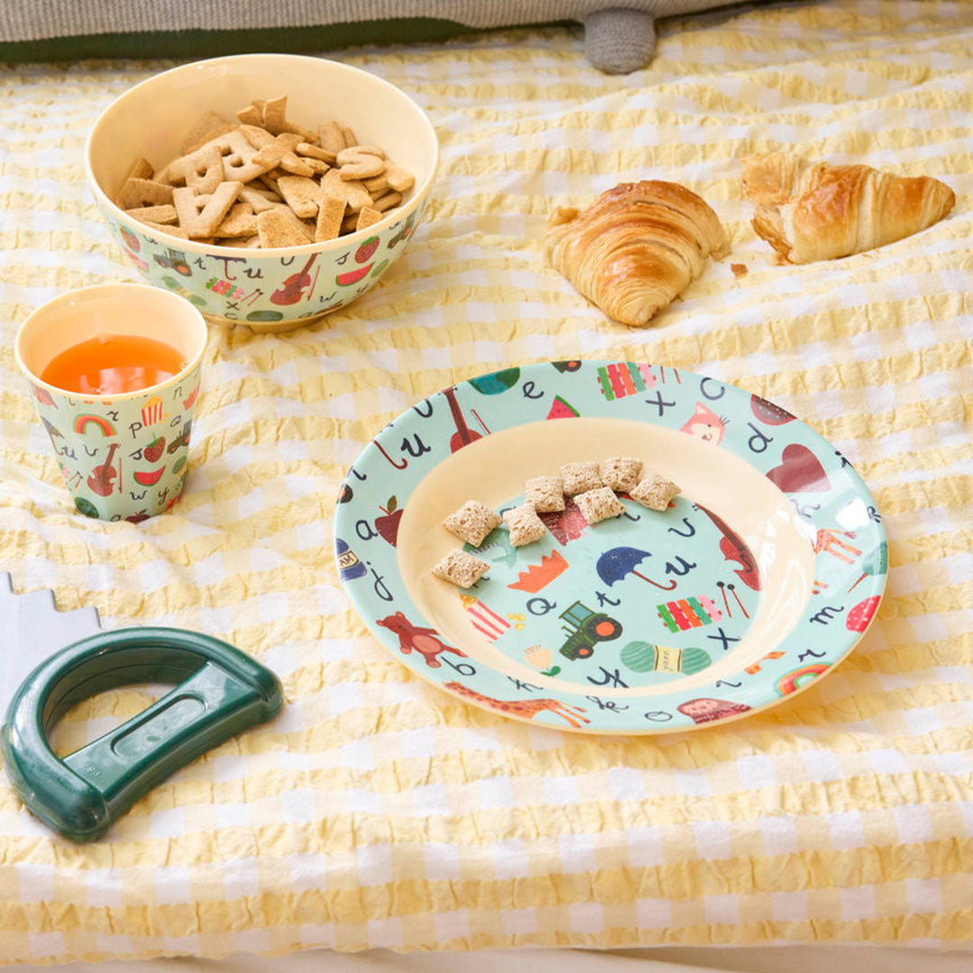 Een kinderbord met ABC-figuren gevuld met ontbijtgranen, naast een bijpassende kom, beker en croissants op een geel geruite deken.