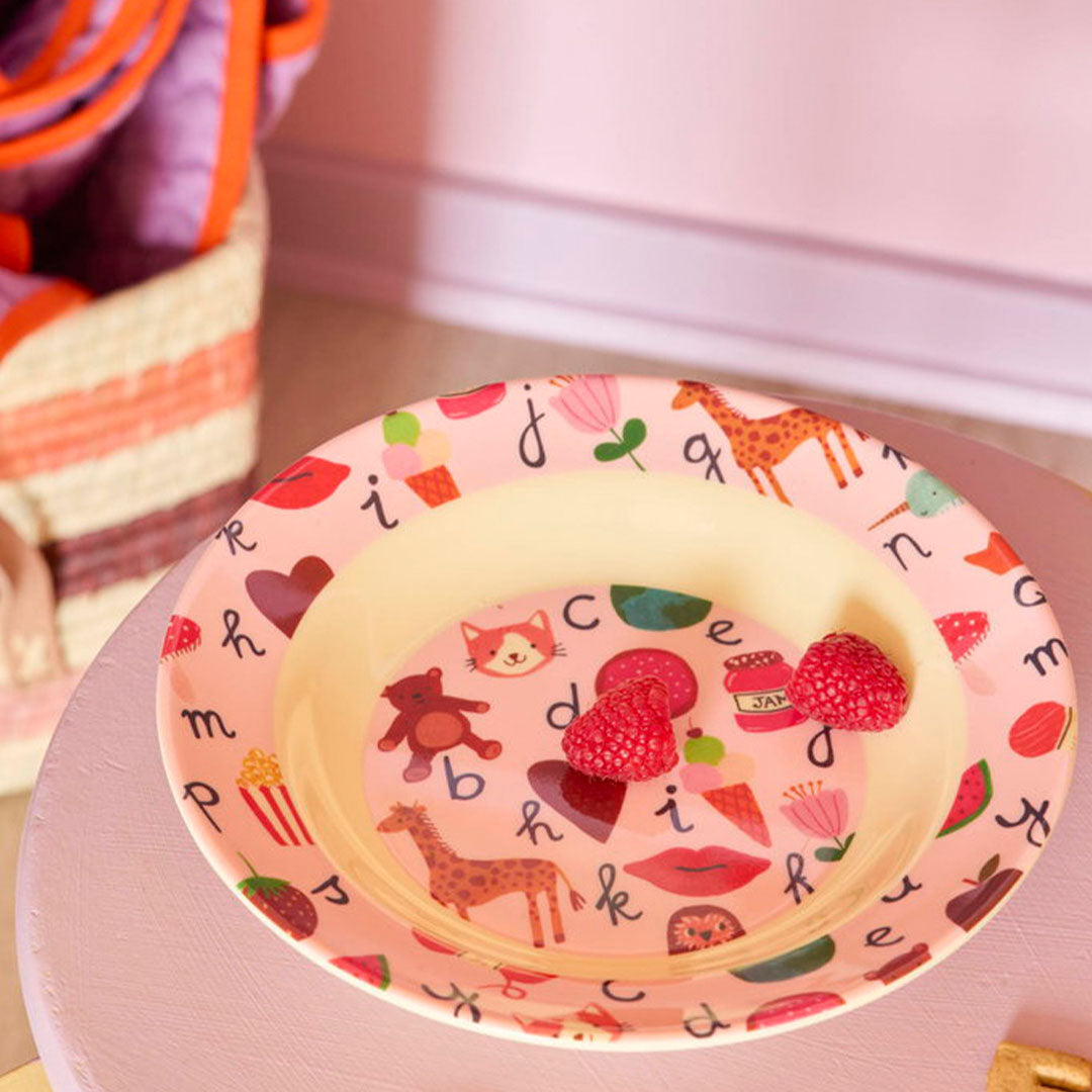 rice kinderbord met roze rand, figuren en alfabetletters, met twee frambozen in het midden op een paarse tafel.