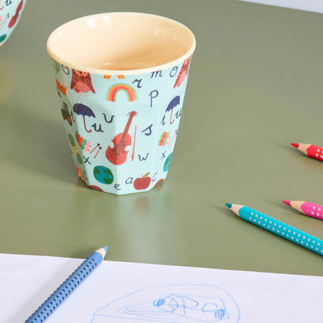 Rice kinderbeker met ABC-patroon op een groene tafel naast kleurpotloden en een tekening.
