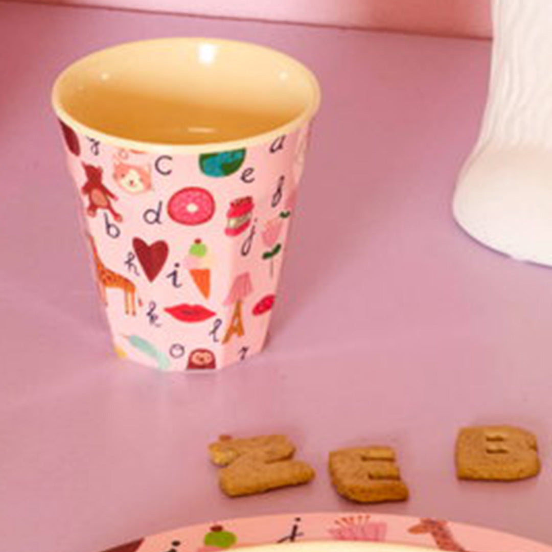 De roze rice kinderbeker met ABC-patroon staat op een paarse ondergrond naast letterkoekjes.