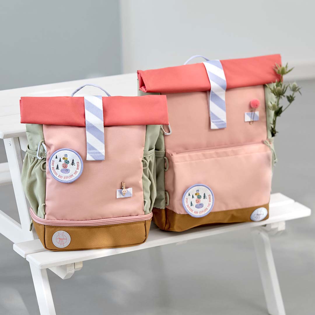 Twee Lässig schoolrugzakken in roze, beige en koraal, een grote en een kleine, staan op een witte bank.