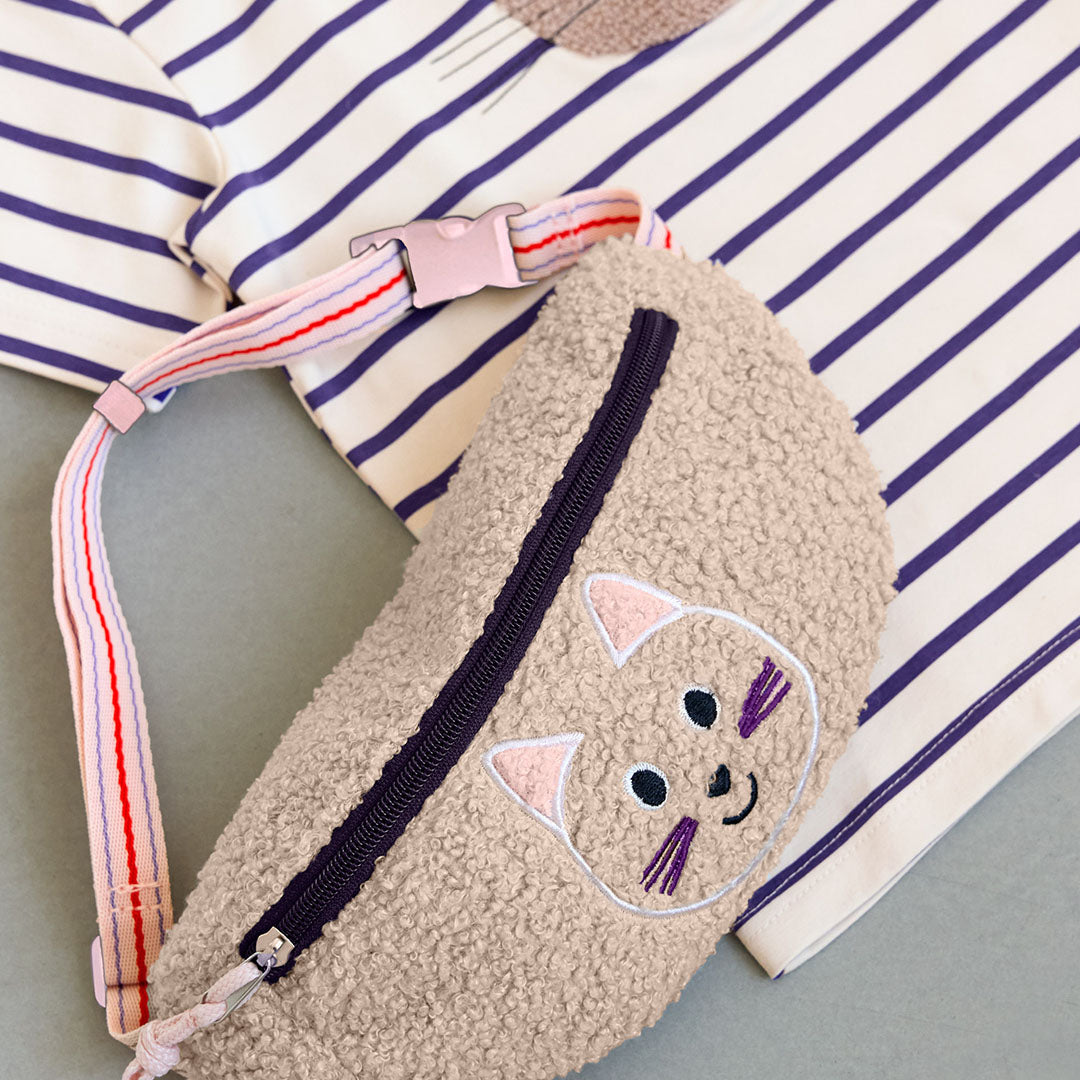 De beige katten heuptas ligt op een grijze ondergrond naast een gestreept shirt.