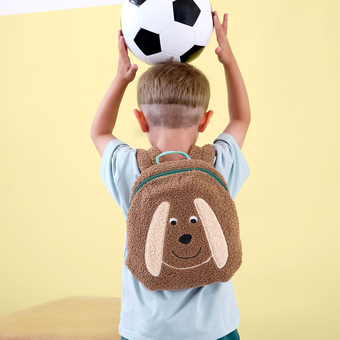 Een jongen draagt de Lässig peuterrugzak met hondenontwerp en houdt een voetbal boven zijn hoofd.