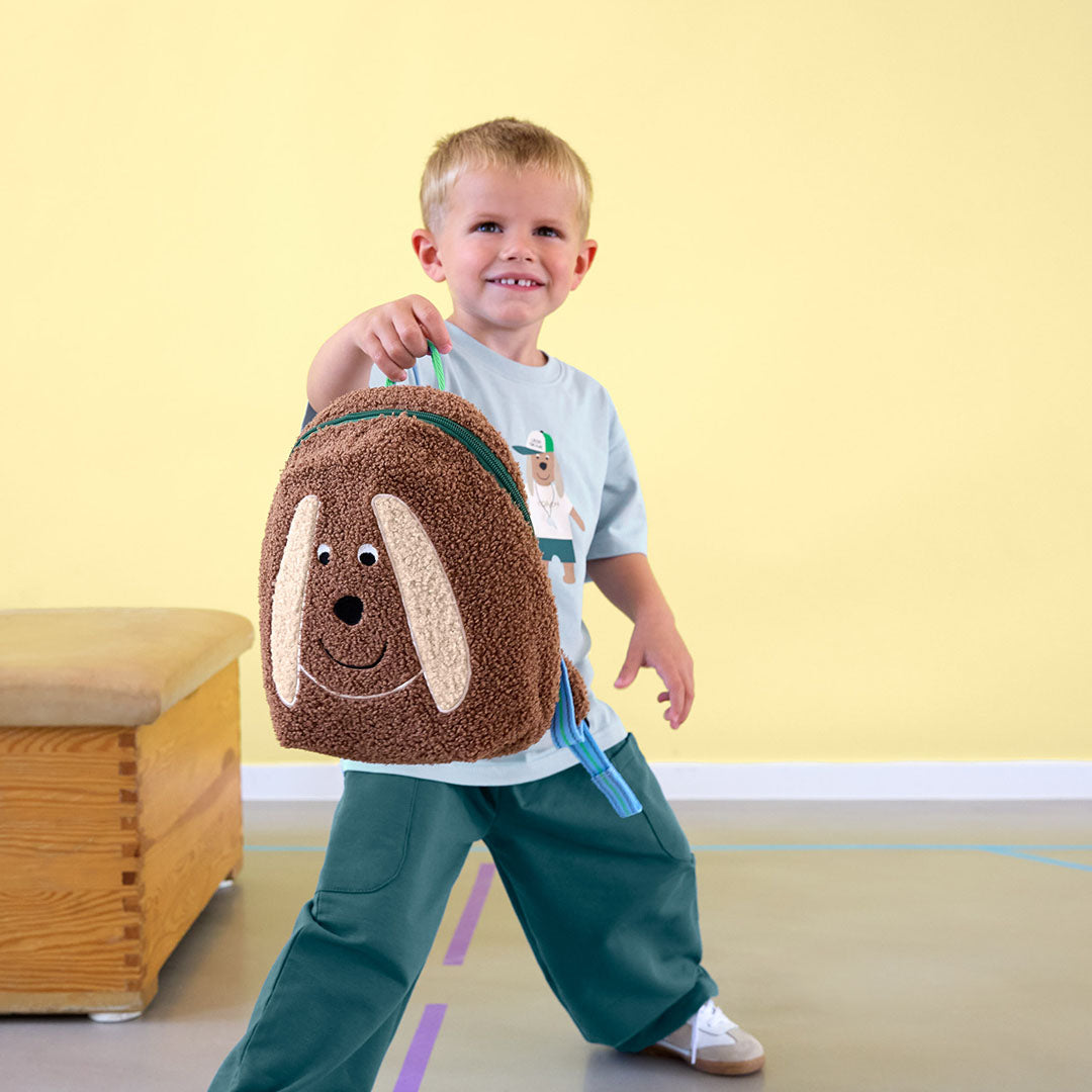 Een lachende jongen houdt de Lässig peuterrugzak met hondenontwerp vast.
