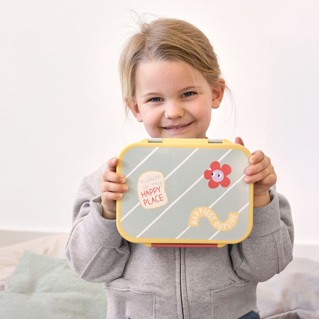 Een lachend kind houdt de Lässig bento-broodtrommel voor kinderen vast, groen met gestreepte deksel, bloem en tekst.