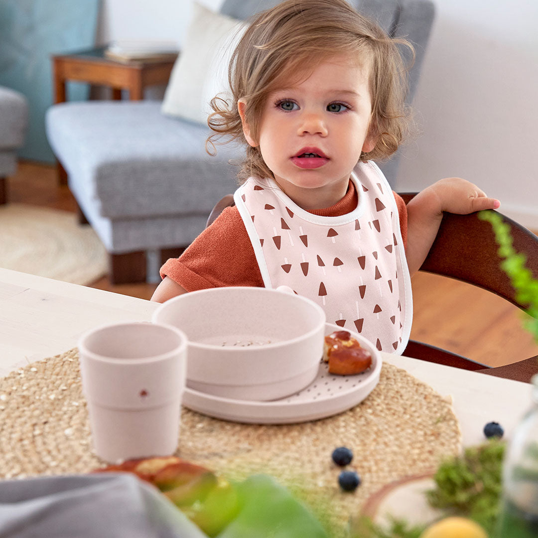 Een peuter met een roze slabbetje zit aan tafel met de lässig beige serviesset.